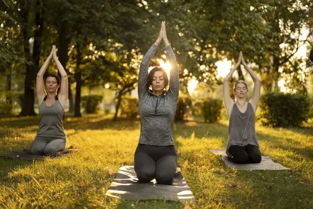 Grupo praticando yoga em um ambiente natural. Três pessoas em um parque praticando ioga com as mãos acima da cabeça, iluminadas pelo sol que filtra pelas árvores.
