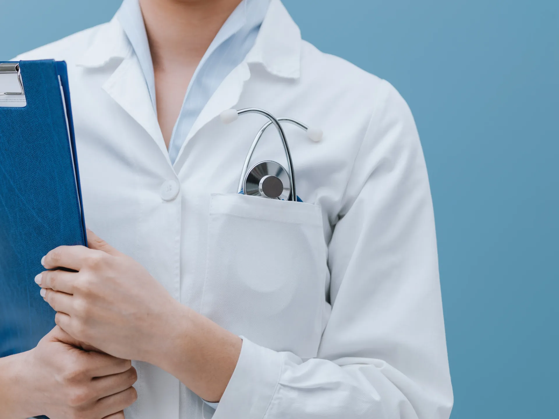 Médico de jaleco branco com estetoscópio no bolso, segurando uma prancheta azul contra um fundo azul claro.