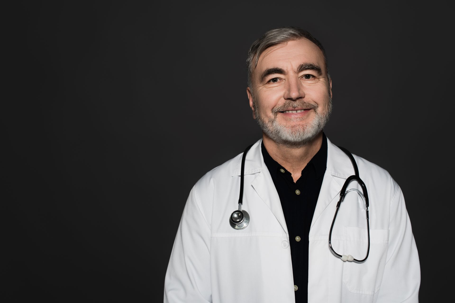Médico de jaleco branco e estetoscópio sorri para a câmera contra um fundo cinza.