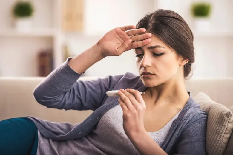 incidência da dengue clássica (CID A90) no Brasil. Mulher deitada no sofá, verificando a temperatura com um termômetro, com expressão preocupada.