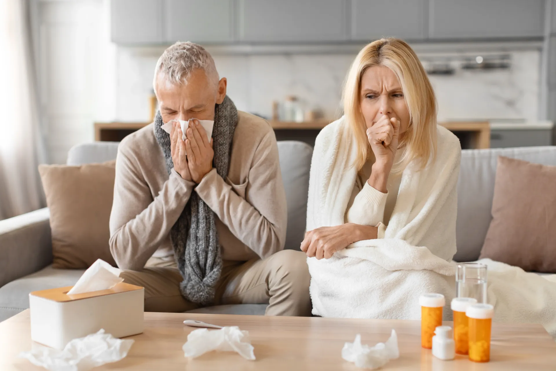 Paciente com gripe – CID J11 sintomas Casal sentado no sofá, doente, com tosse e lenços de papel. Frascos de remédio sobre a mesa.