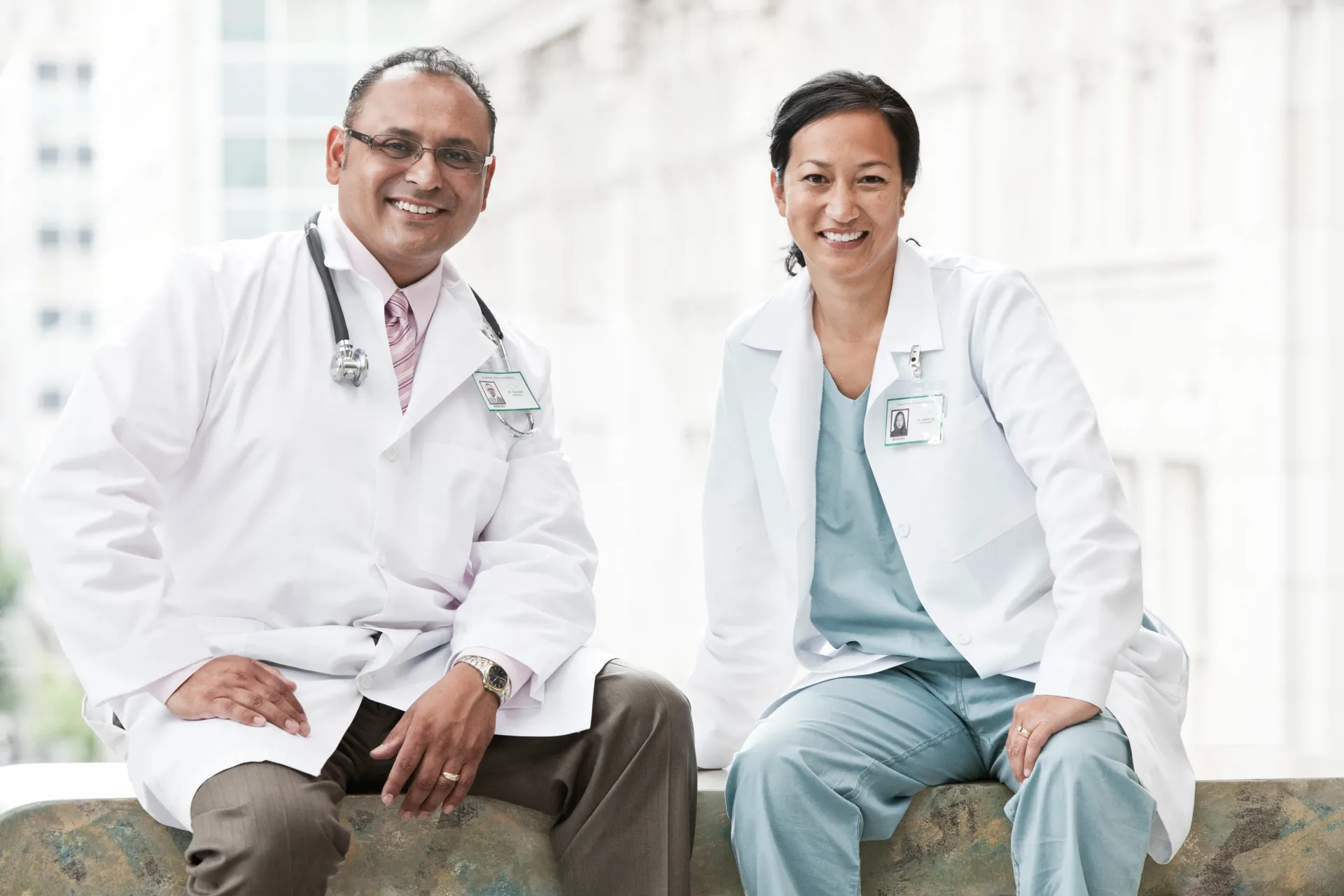 Dois médicos de jaleco branco sorriem, sentados do lado de fora.