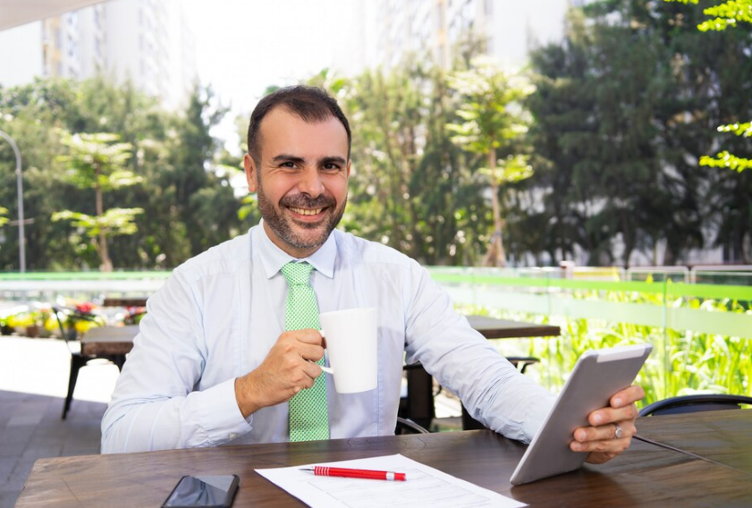 Homem de camisa azul-clara e gravata verde, sorrindo, segurando uma caneca e um tablet em uma mesa de café ao ar livre.