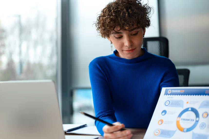 Uma mulher de suéter azul analisa um gráfico financeiro, caneta na mão, sentada à mesa com um laptop.