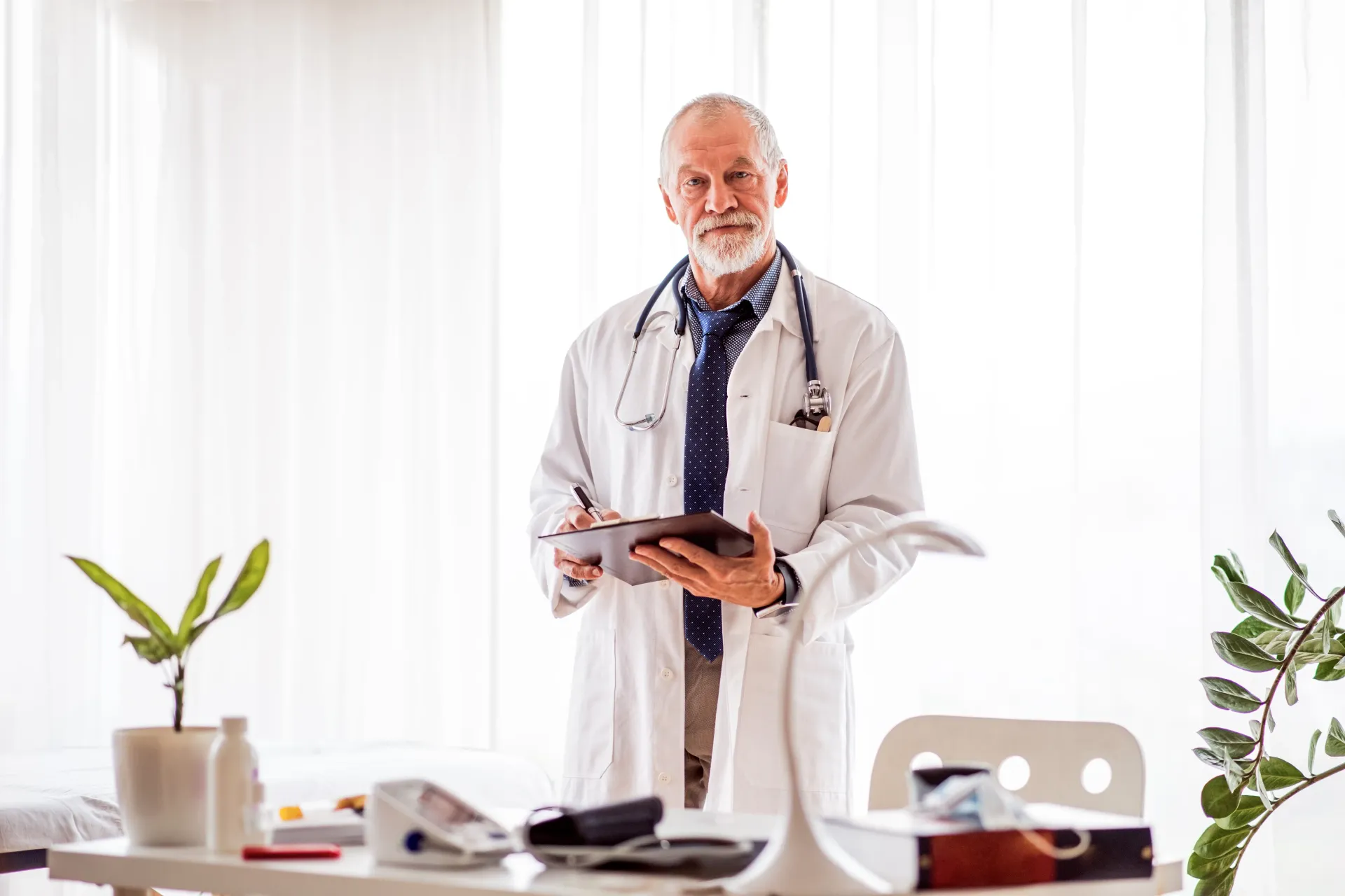 Médico de jaleco branco, estetoscópio, segurando um tablet, em pé no consultório, olhando para a câmera.