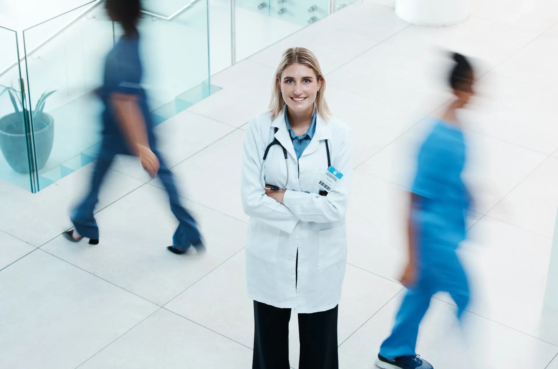 Médico de jaleco branco, braços cruzados, sorrindo no corredor do hospital. Funcionários da área médica passam desfocados.