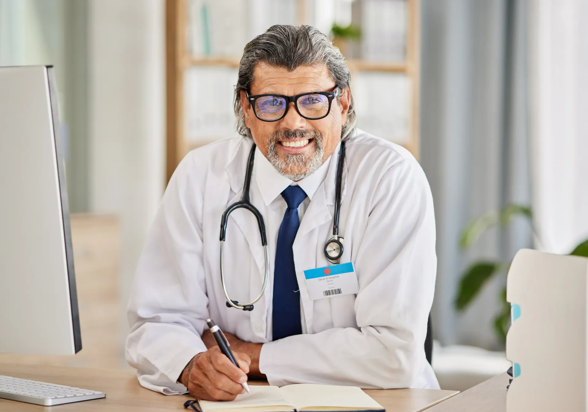 Médico sentado à mesa, sorrindo, usando óculos e estetoscópio, escrevendo em um bloco de notas.