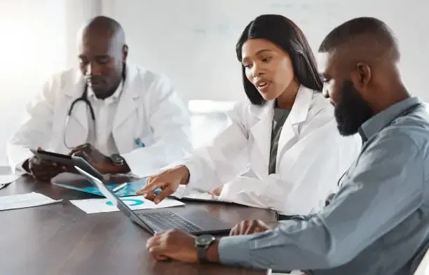 Médico analisando relatório financeiro da clínica Três profissionais da área médica colaborando em torno de um laptop e documentos em uma sala de reuniões.