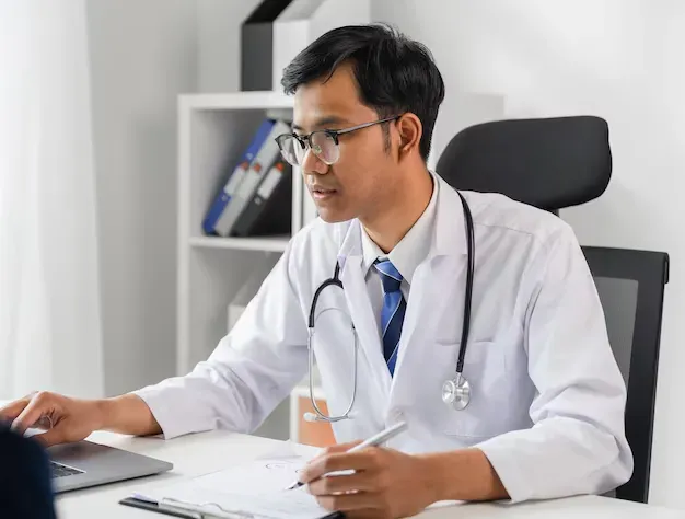 sistema de gestão de repasse e comissionamento da App Health Médico de jaleco branco, estetoscópio, trabalhando em uma mesa com um laptop, escrevendo.