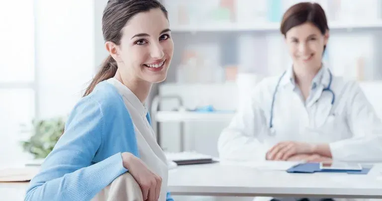 Profissional configurando permissões de acesso em software de gestão médica Uma mulher de suéter azul sorri para a câmera em um consultório médico. O médico de jaleco branco sorri ao fundo.