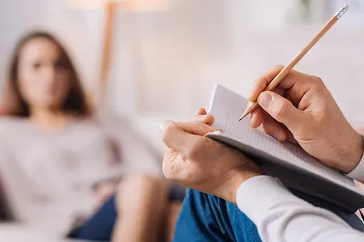 psicólogo explicando o processo de encaminhamento psicológico a um paciente. Terapeuta fazendo anotações durante uma sessão; paciente sentado em foco suave ao fundo.