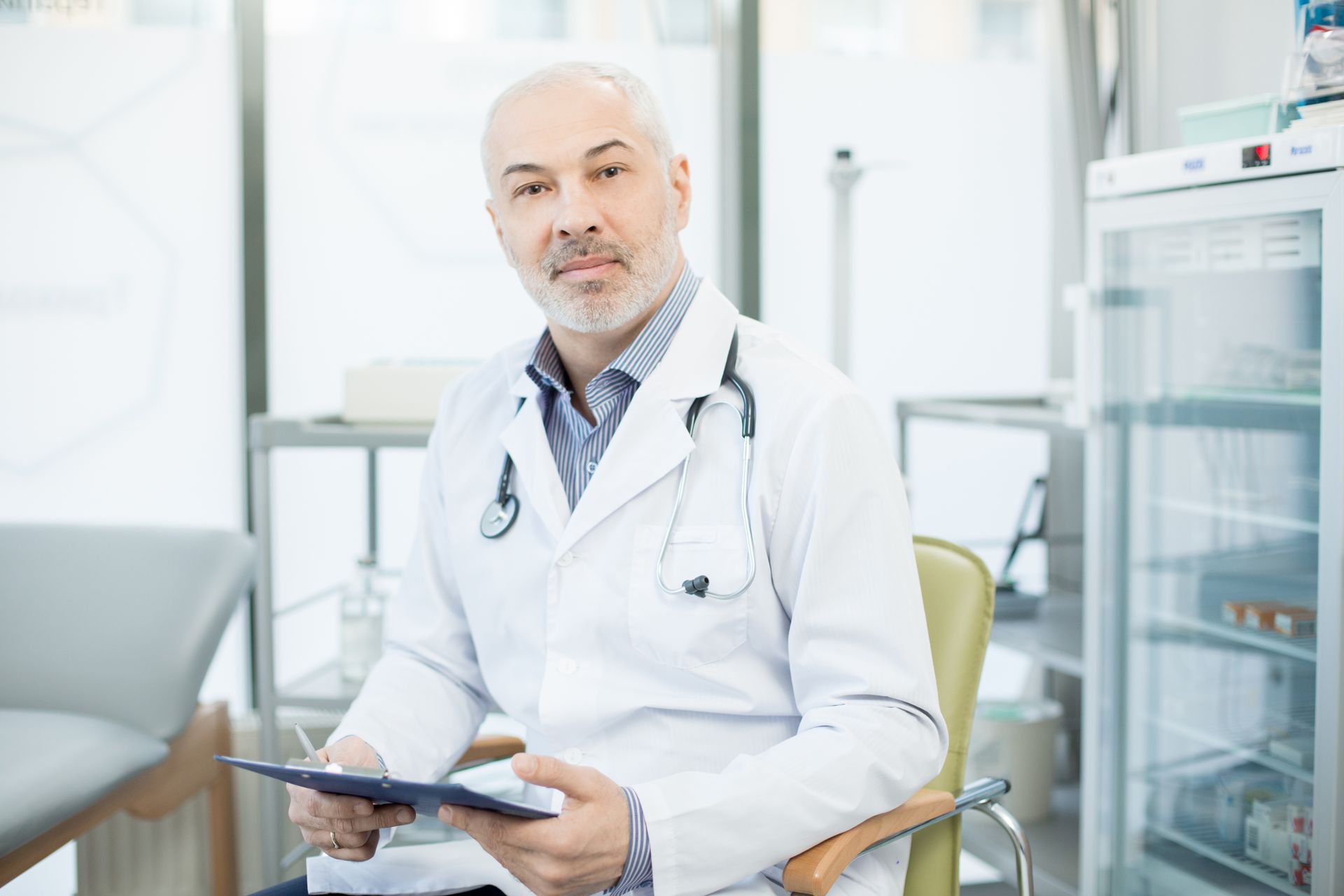 Médico de jaleco branco com estetoscópio, segurando uma prancheta, olhando para a câmera.