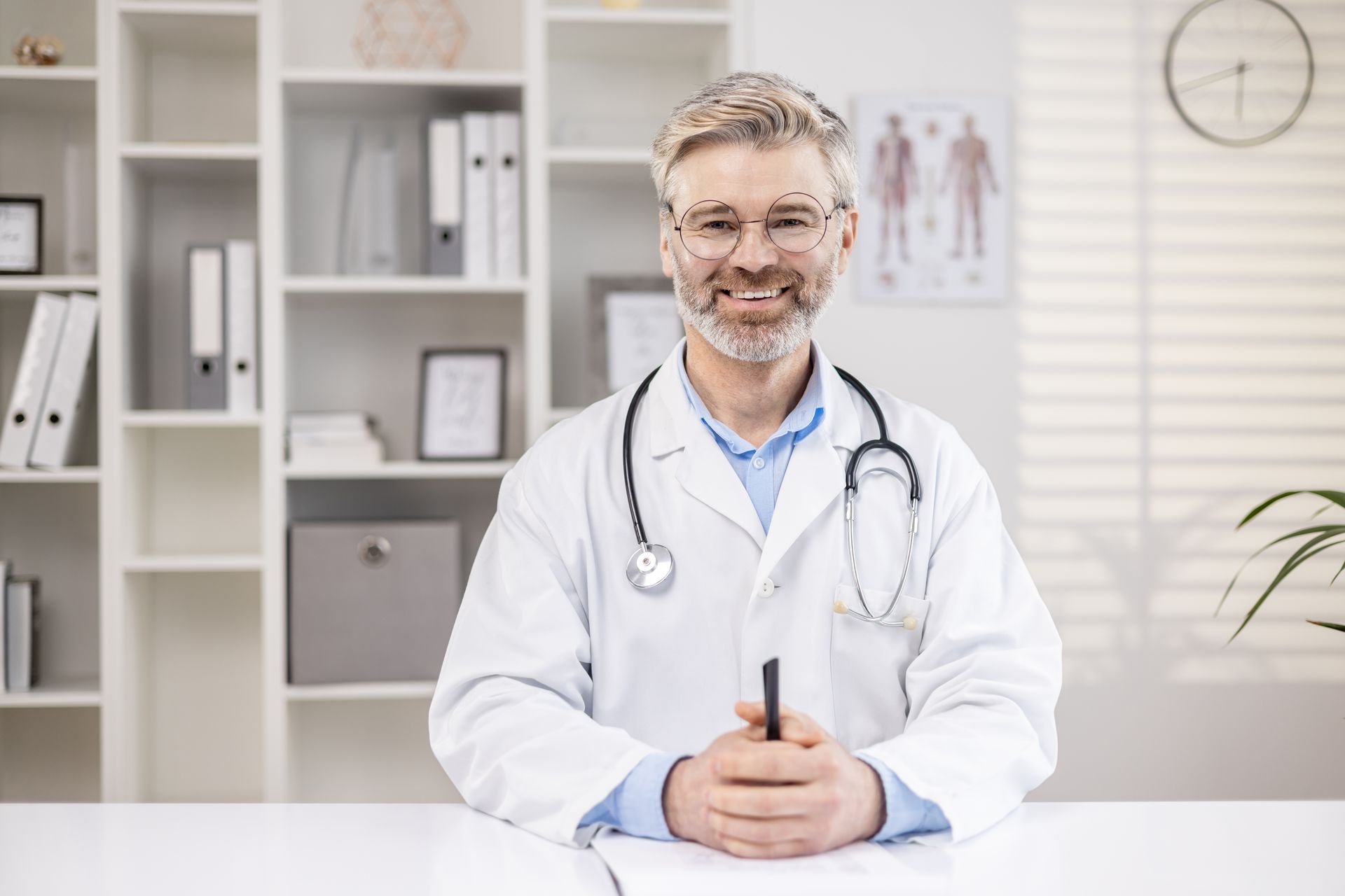 Médico de jaleco branco, sorrindo, com estetoscópio na mesa do consultório médico.