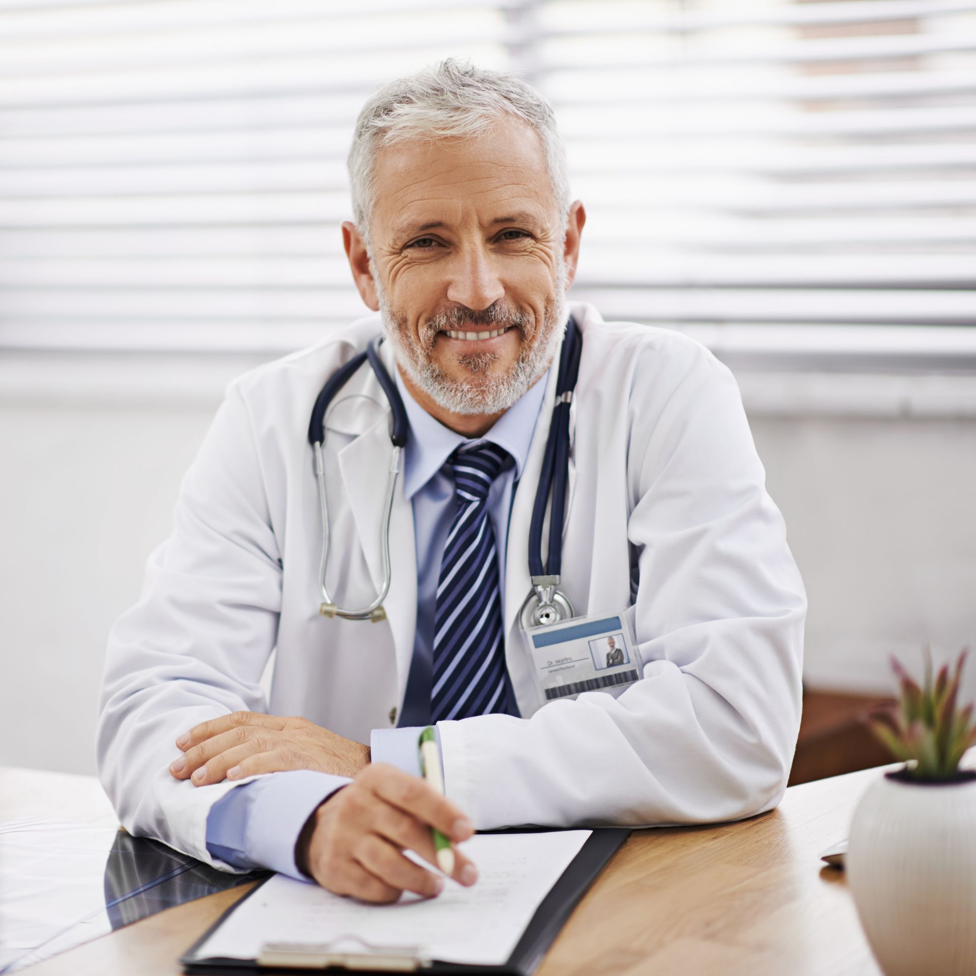 Médico sorrindo, sentado à mesa, vestindo jaleco branco e estetoscópio.