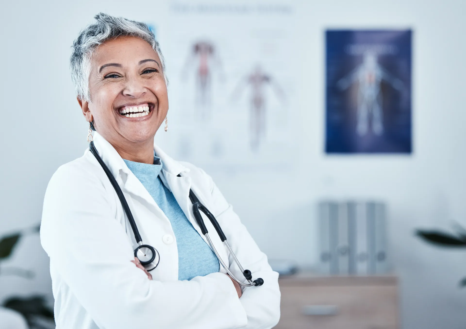 Médica de braços cruzados, sorrindo em um consultório médico, com estetoscópio no pescoço.