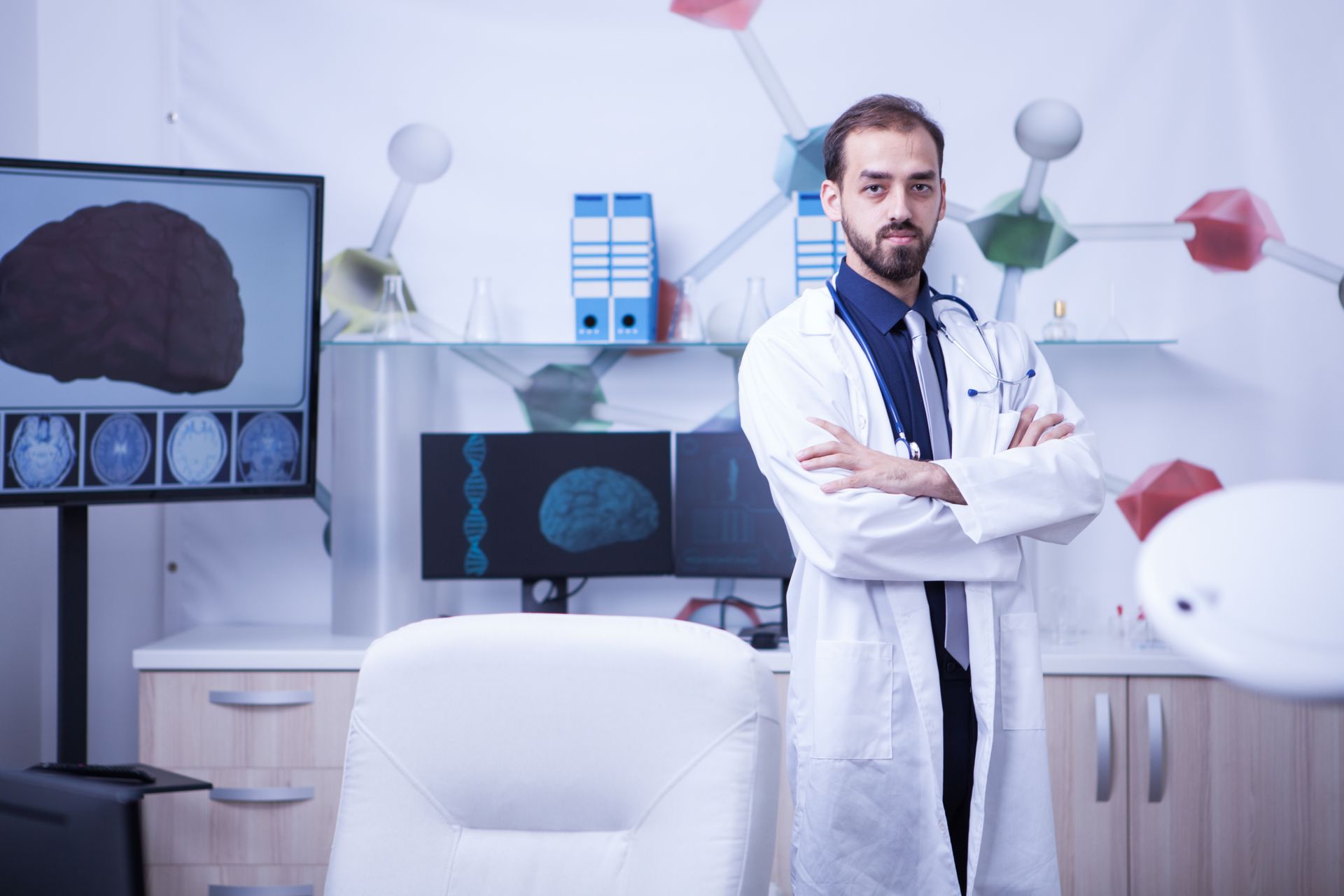 alergista e imunologista Médico de jaleco branco, com os braços cruzados, em pé num consultório com imagens médicas em telas.