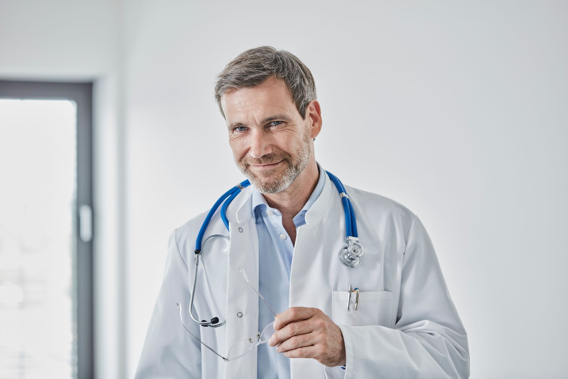 Médico de jaleco branco, segurando óculos, com estetoscópio no pescoço, sorrindo.