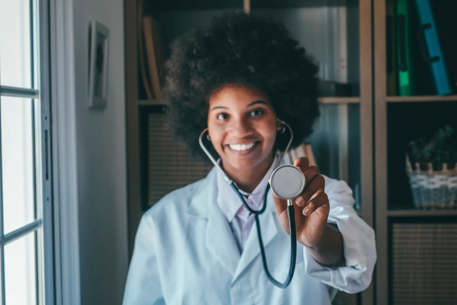Médico segurando um estetoscópio, sorrindo, vestindo um jaleco branco, perto de uma janela e uma estante de livros.