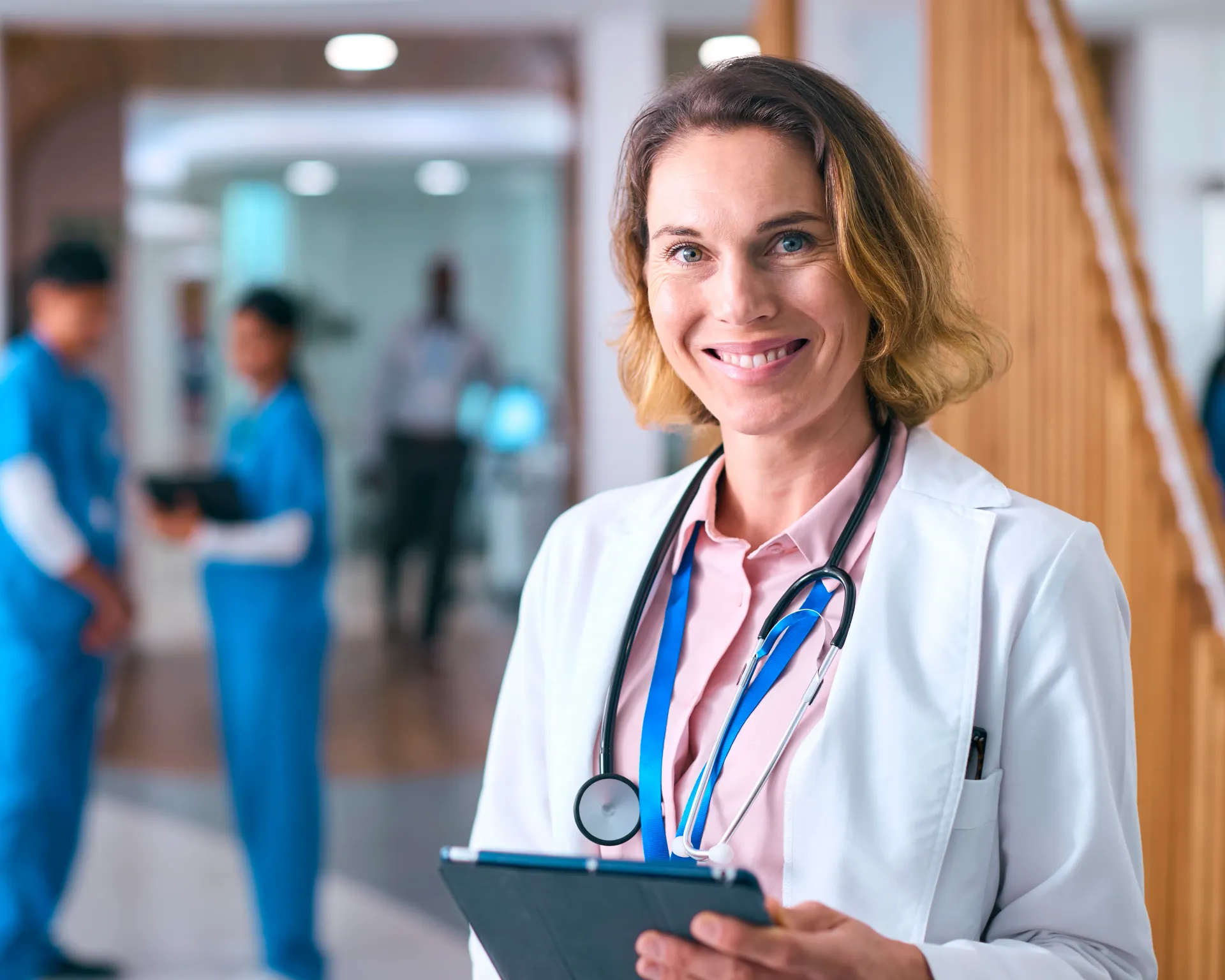 Uma médica sorri para a câmera, vestindo um jaleco e um estetoscópio, segurando um tablet em um ambiente médico.