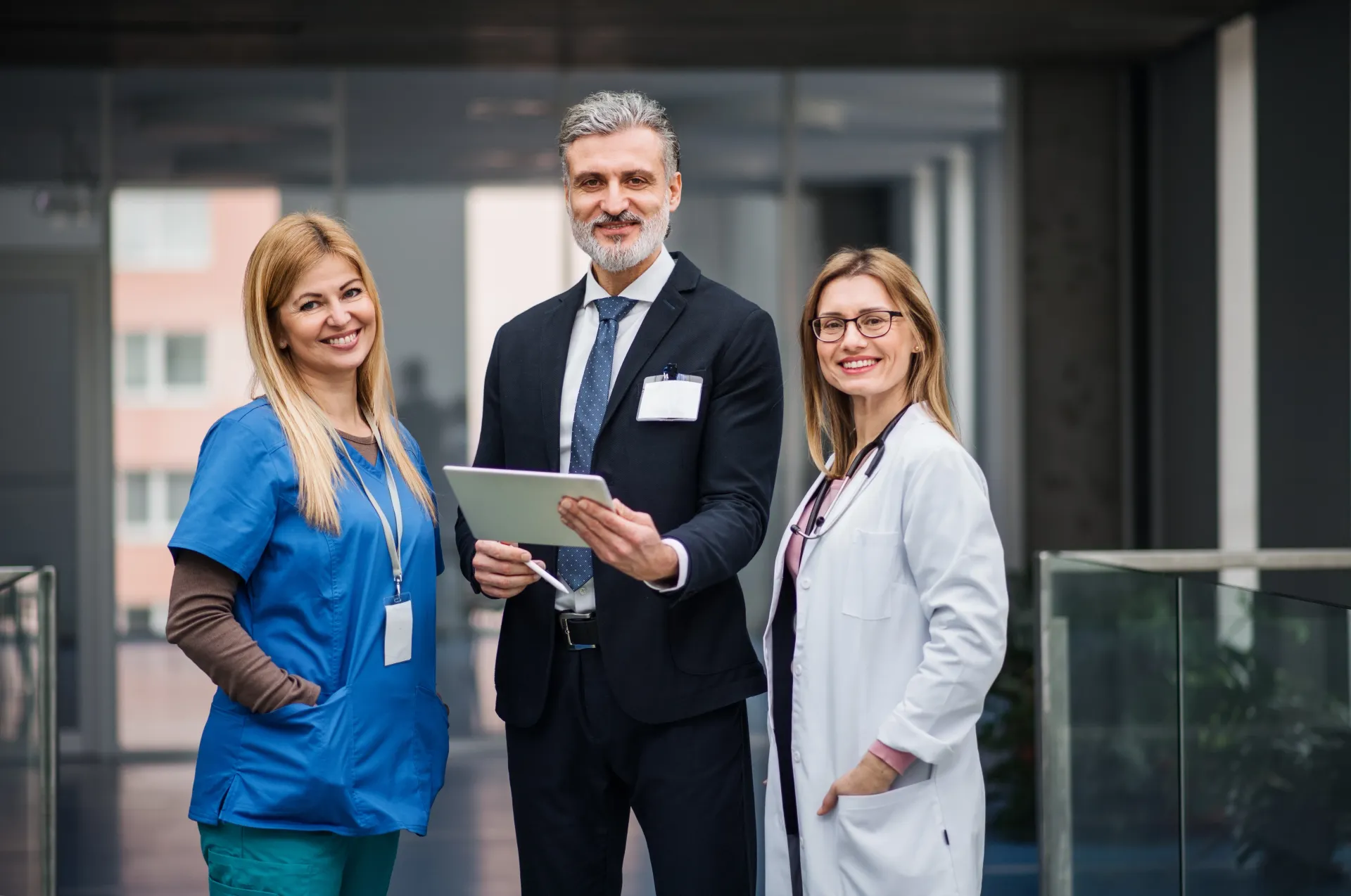 Três pessoas em um corredor moderno. Um homem de terno segura um tablet, ladeado por uma enfermeira de uniforme azul e um médico de jaleco; todos estão sorrindo.