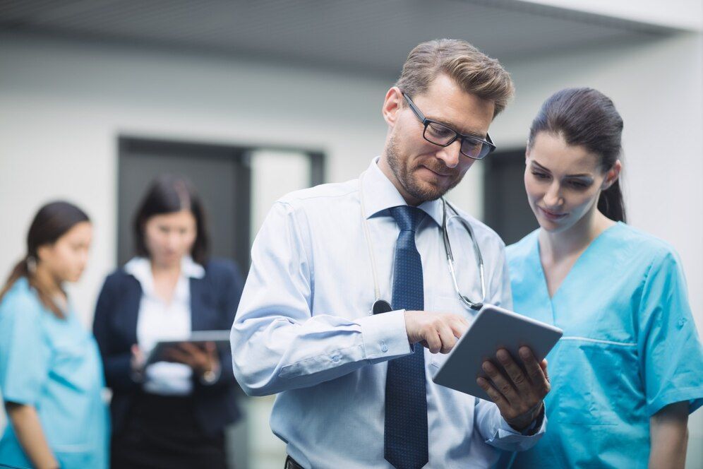 Médico e enfermeira analisando o tablet; outros funcionários ao fundo.