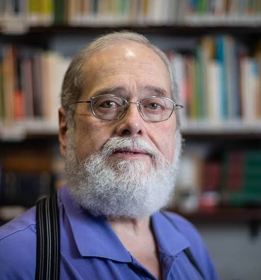 Homem de óculos e barba branca, vestindo uma camisa roxa, em frente a uma estante de livros.