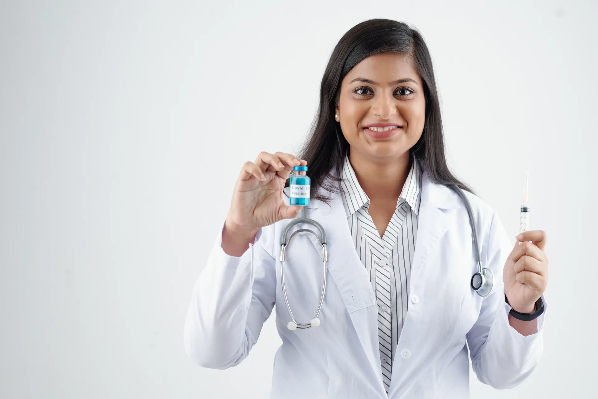 Médico segurando frasco e seringa de vacina, sorrindo, vestindo jaleco e estetoscópio.
