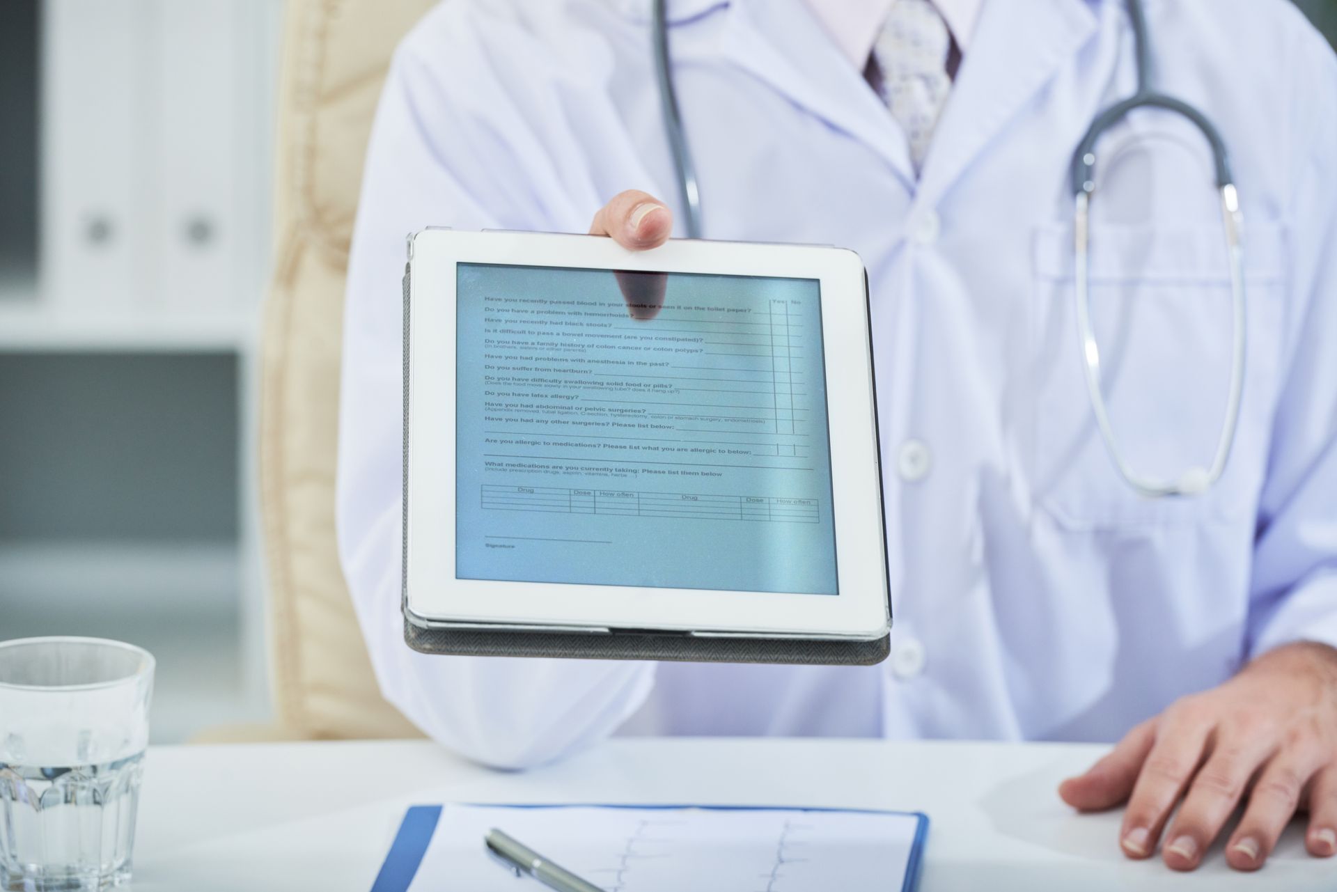 Médico segurando um tablet com informações médicas, estetoscópio no pescoço, sentado à mesa.