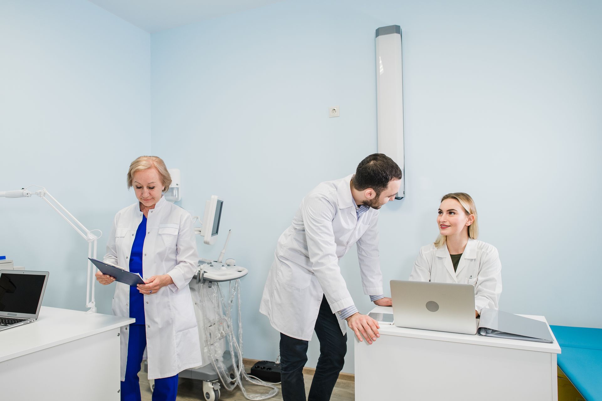 Três profissionais da área médica, trajando jalecos brancos, examinam prontuários de pacientes e um computador portátil em uma clínica.