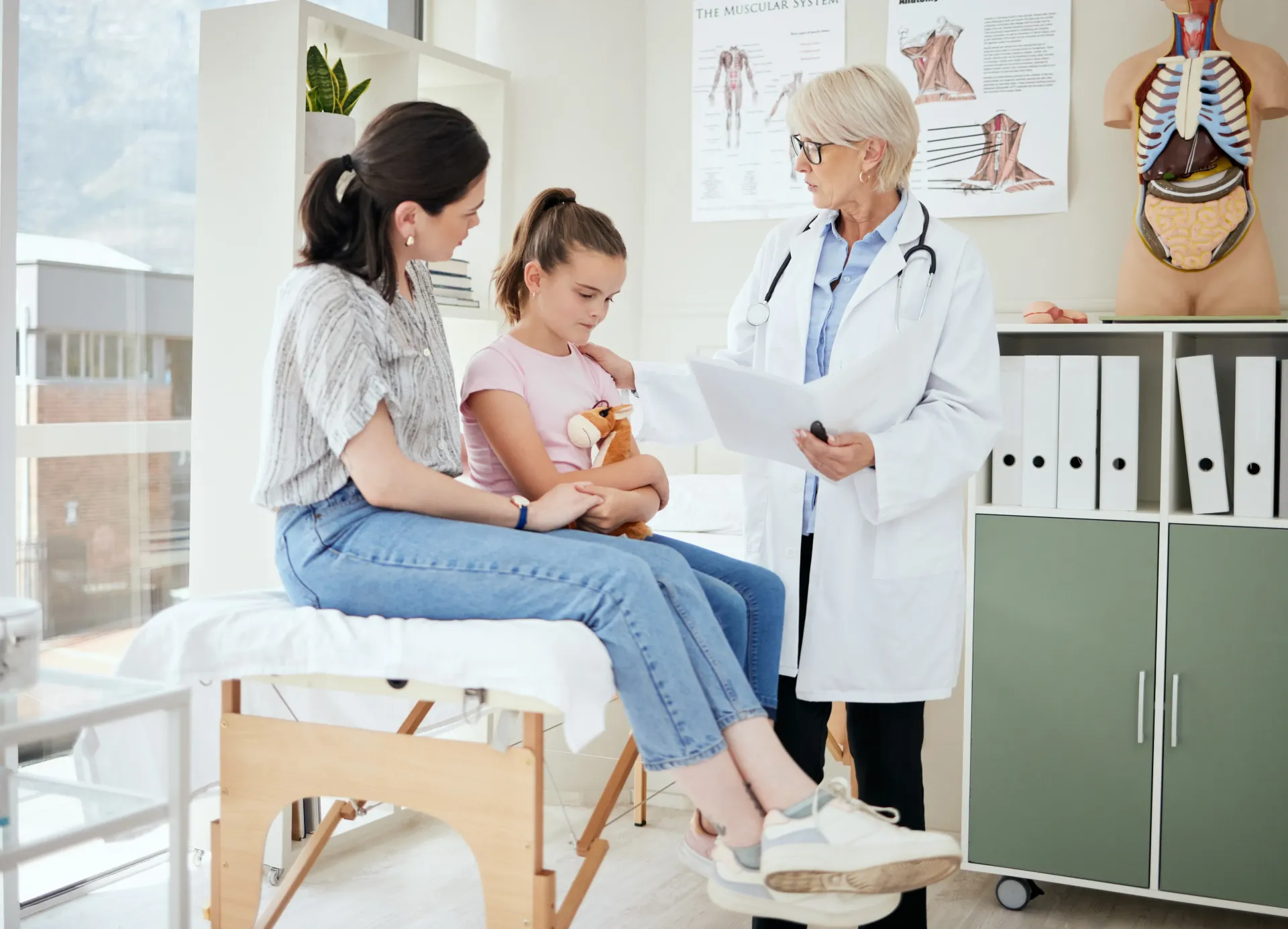 Mãe e filho no consultório médico. O médico está revisando documentos. A criança segura um brinquedo.