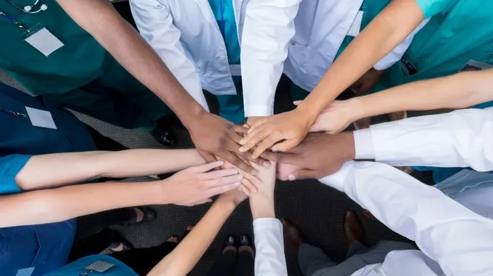 Mãos de profissionais da saúde reunidas em círculo, simbolizando trabalho em equipe e apoio mútuo.