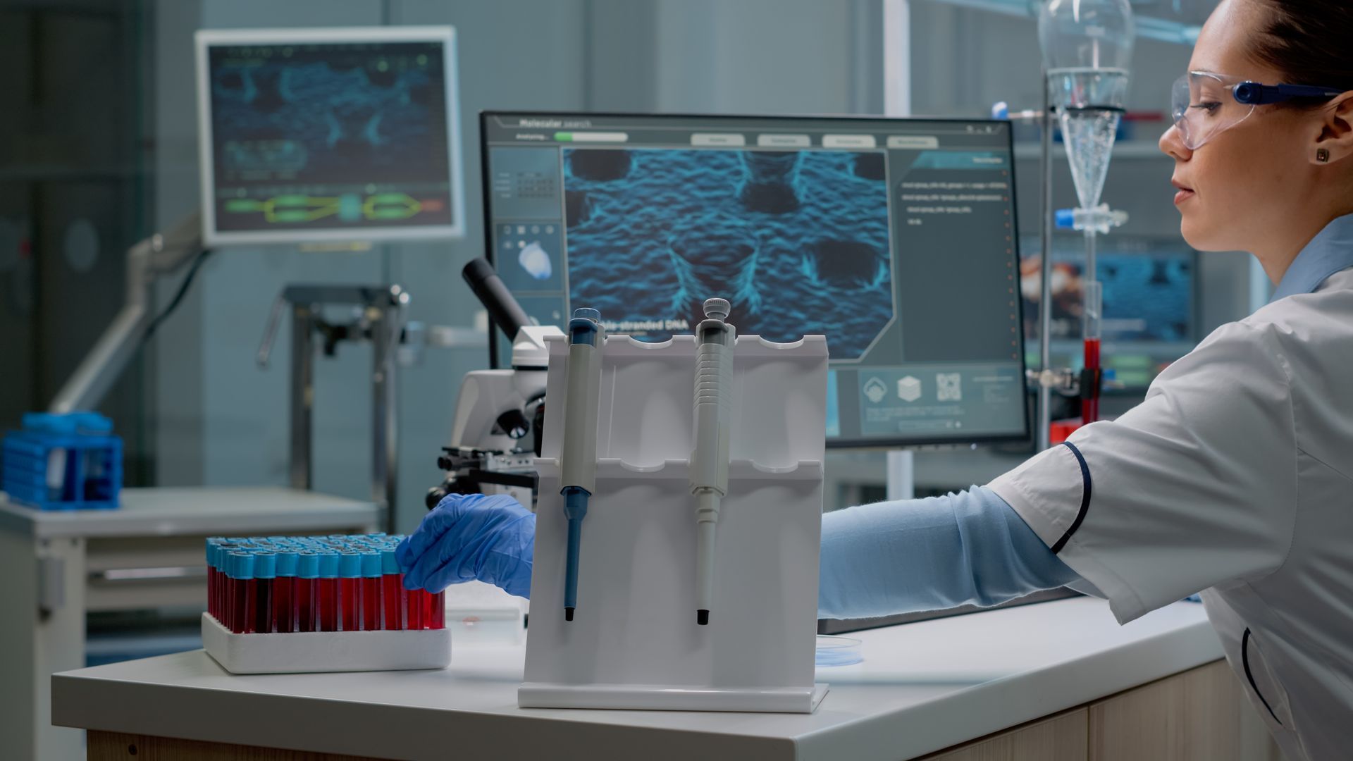 Técnico de laboratório com luvas, utiliza pipeta, analisa computador com imagens azuis; ambiente médico.