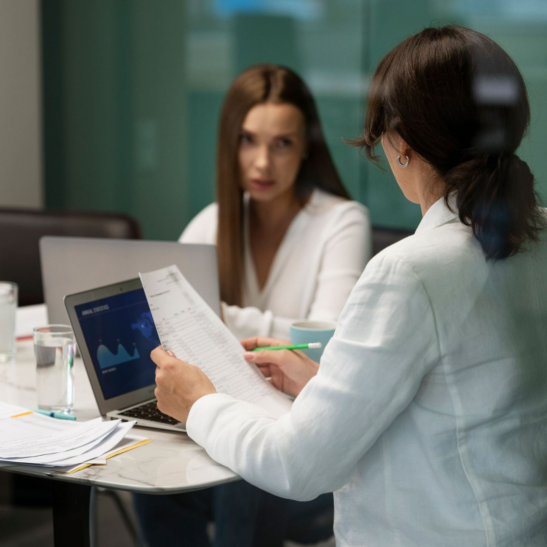 Duas mulheres revisando documentos, uma delas usando um laptop, em um ambiente de escritório.