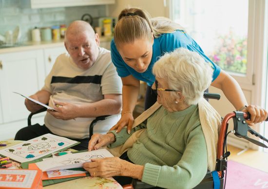 Woman assisting elderly couple with crafts at a table, sunny room.