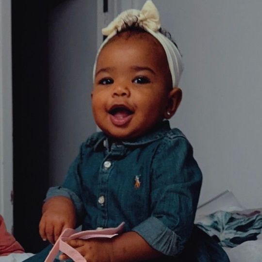 A baby wearing a headband and a denim dress is smiling