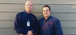 Two individuals in matching dark blue collared work shirts stand side-by-side against a light brown horizontal siding wall.