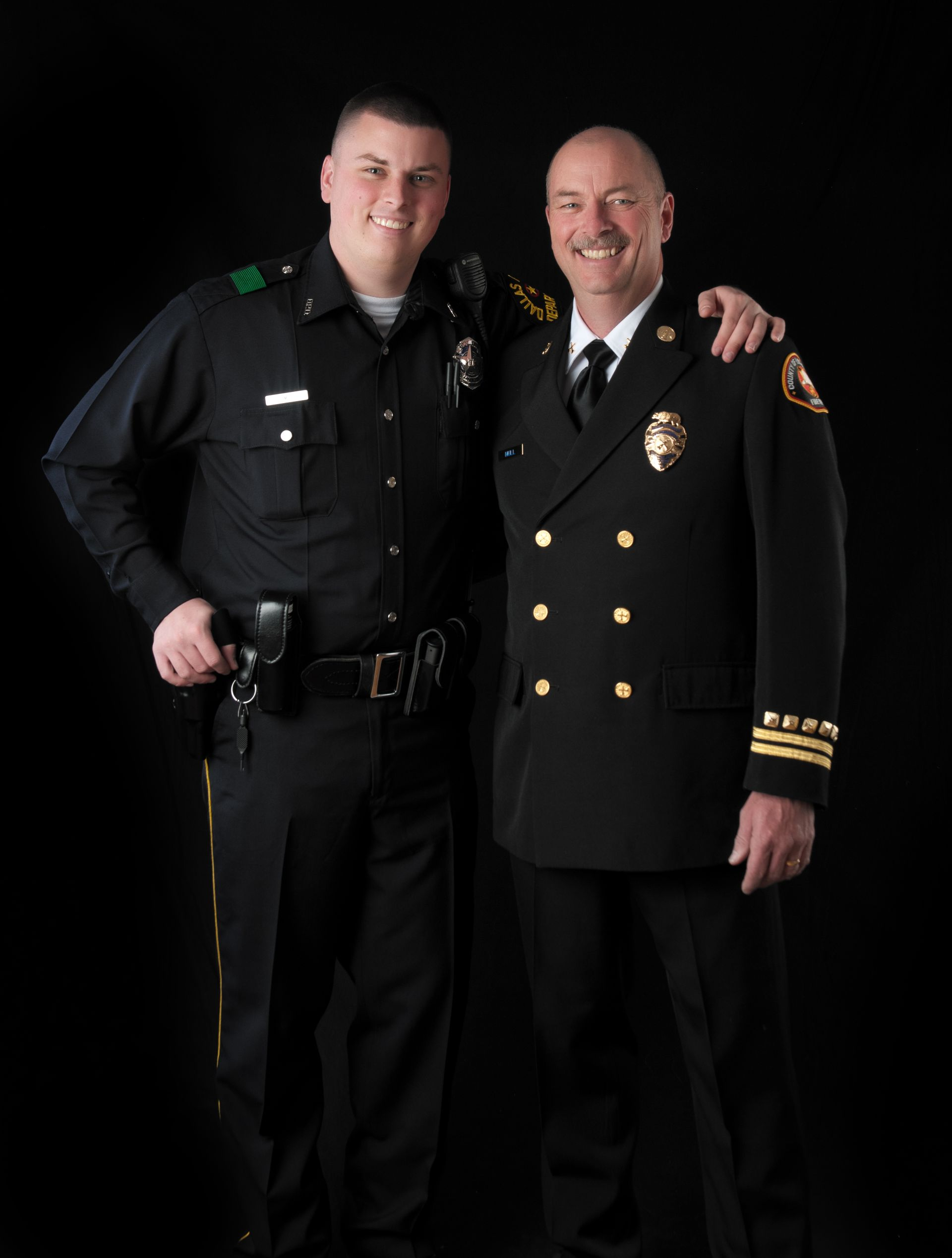 Police officer and firefighter standing together, smiling, against a black background.
