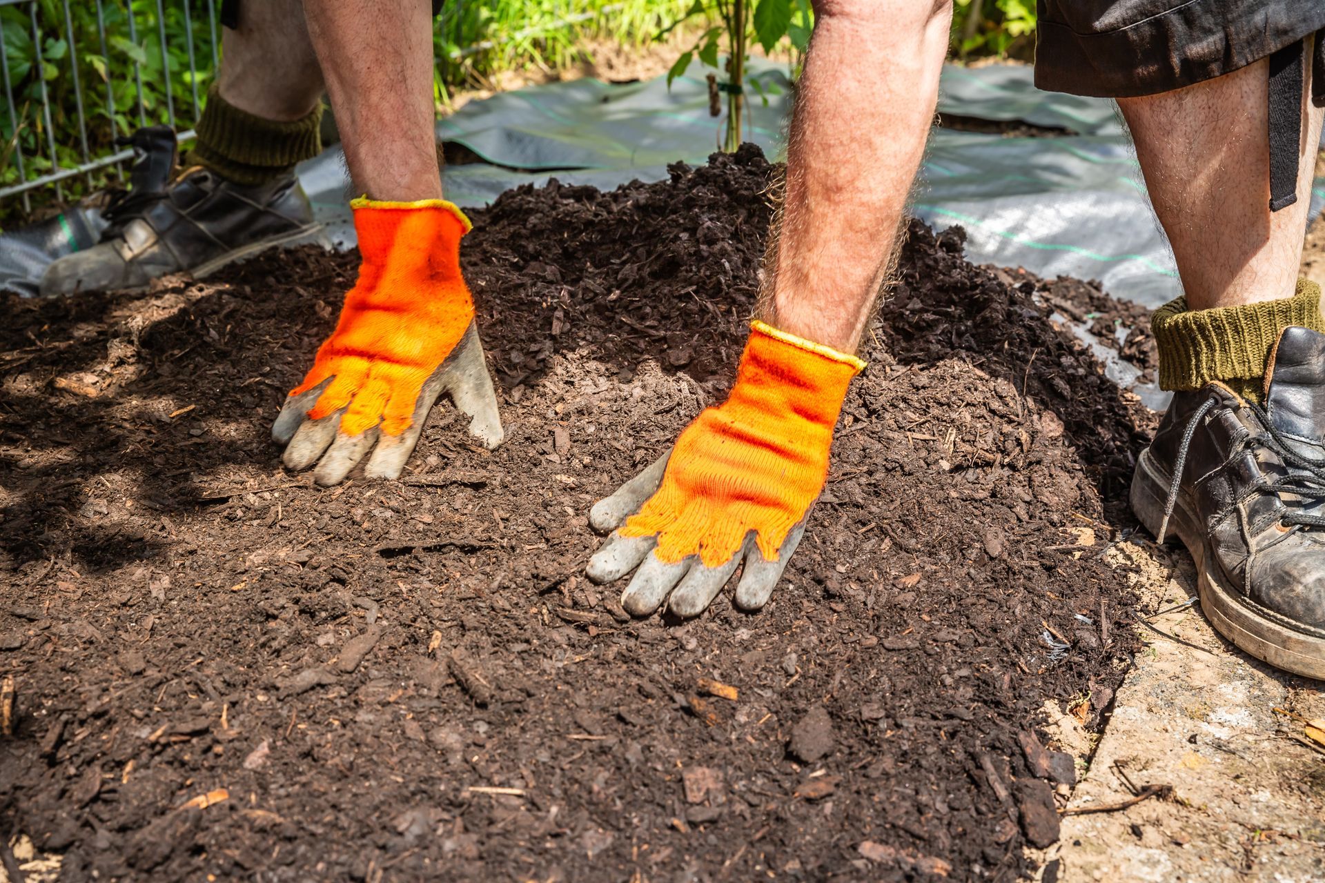 Gloved hands spreading dark soil over a garden bed beside boots and a tarp