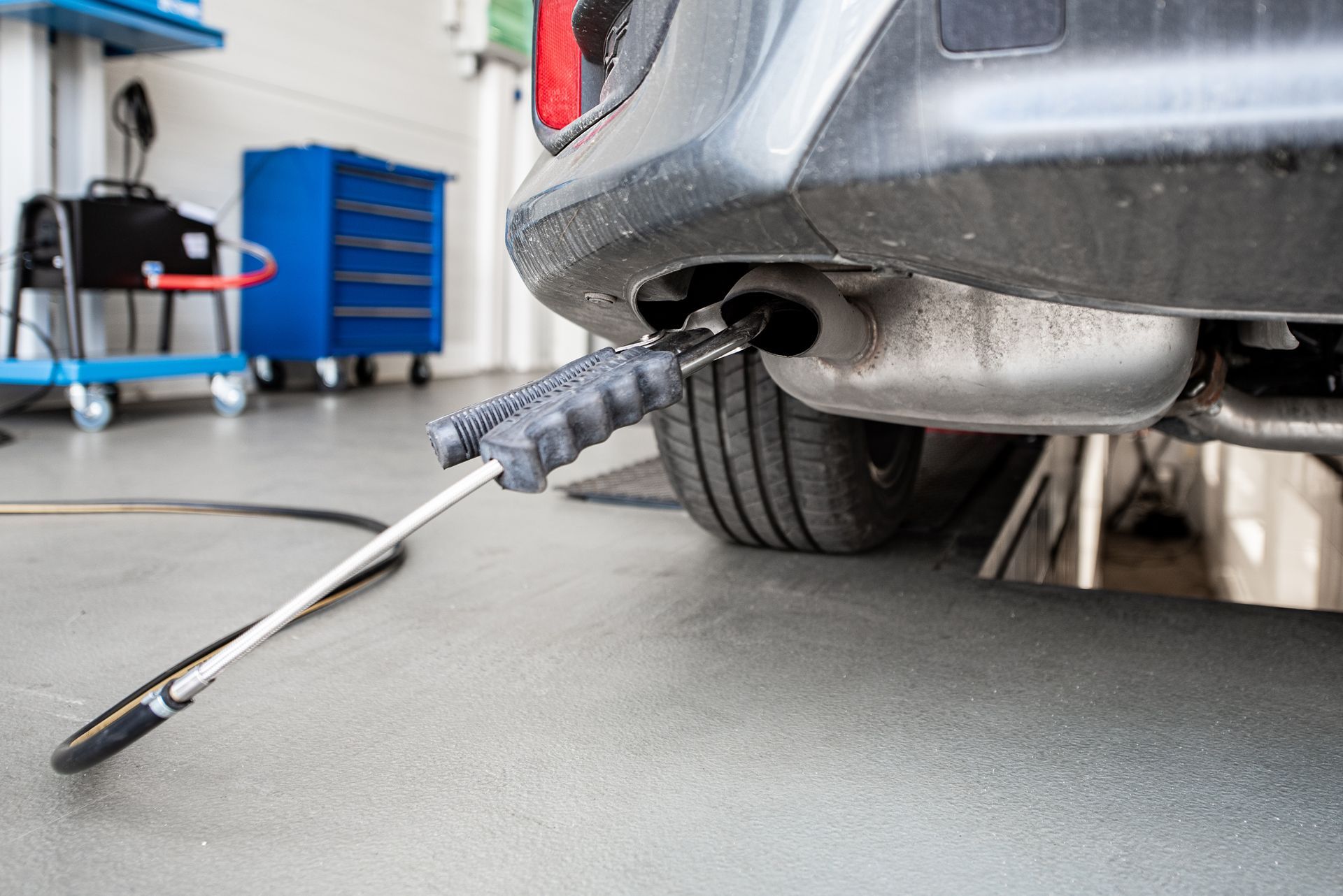 Exhaust emissions test: Probe in car's tailpipe, grey car, mechanic's garage setting.