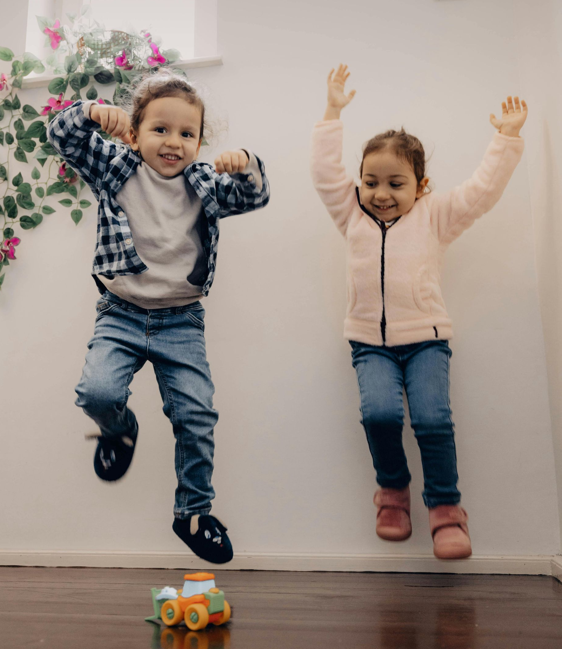 Two children jumping indoors with arms raised, smiling beside a small toy car on the floor.