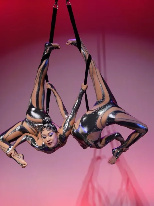 Two Milanus Circus aerialists in sparkling silver costumes hanging from black straps performing synchronized backbends.