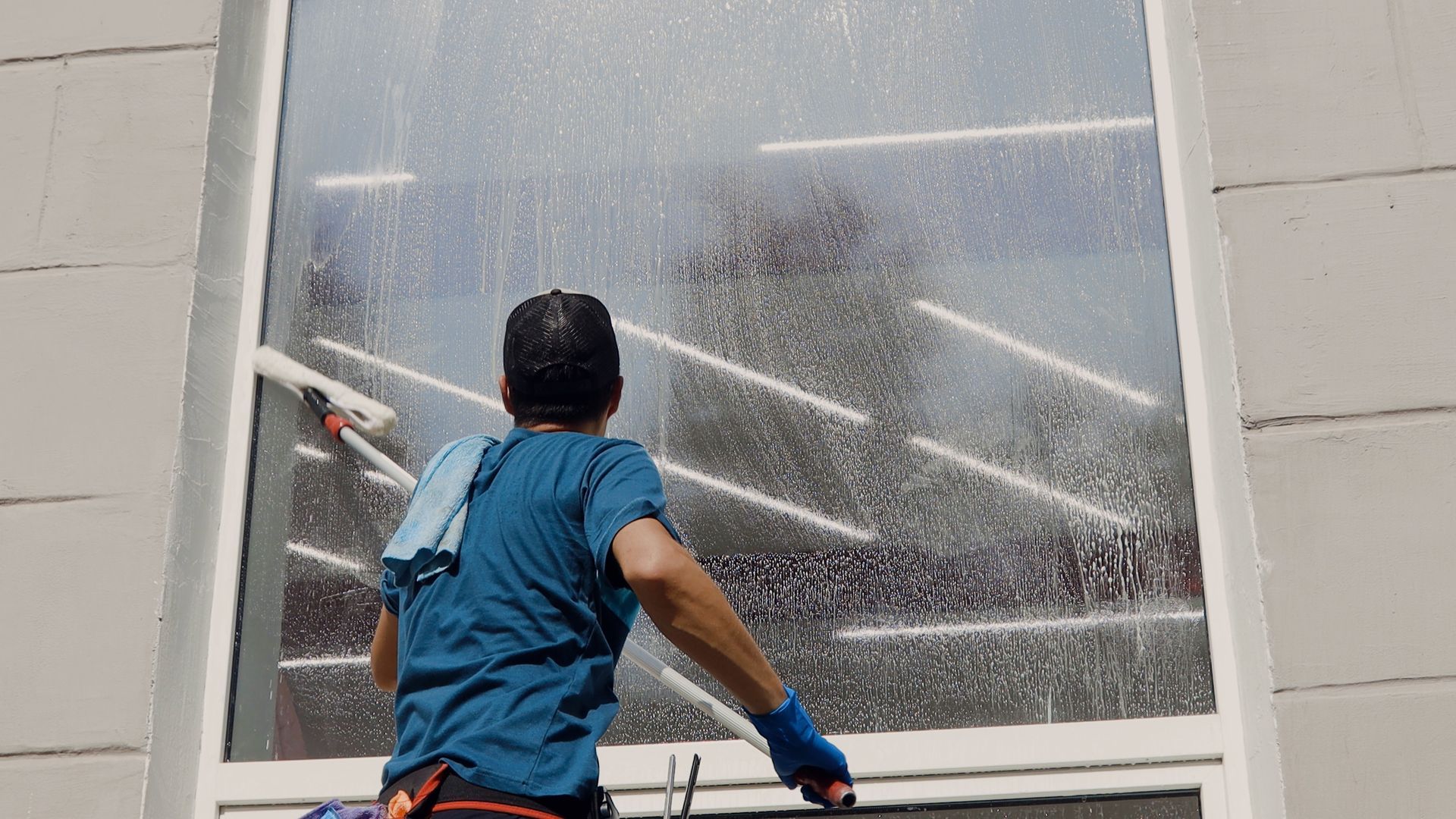 Worker spray-washing a large window with a hose, water splashing across the glass.