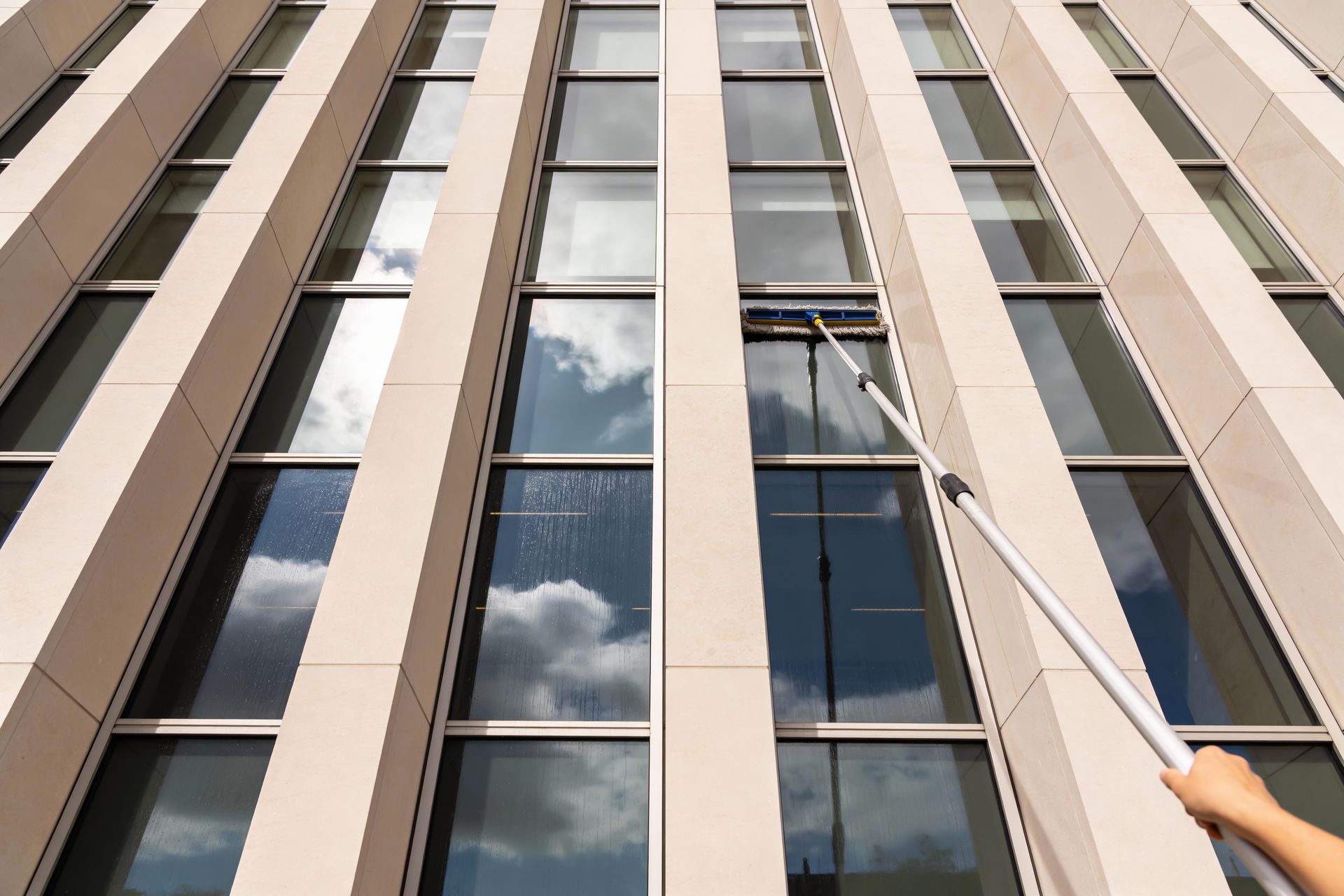 Person cleaning tall building windows with a long-handled squeegee, reflecting clouds in glass