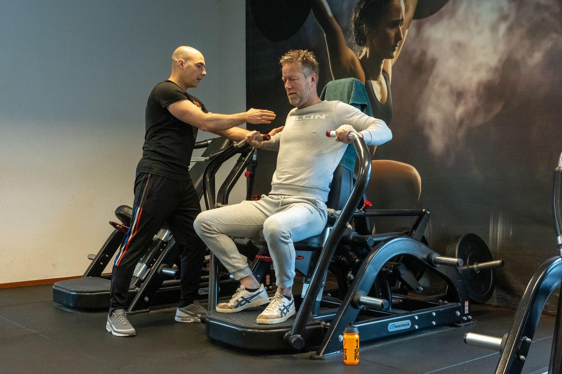 Een man zit op een machine in een sportschool terwijl een andere man achter hem staat.