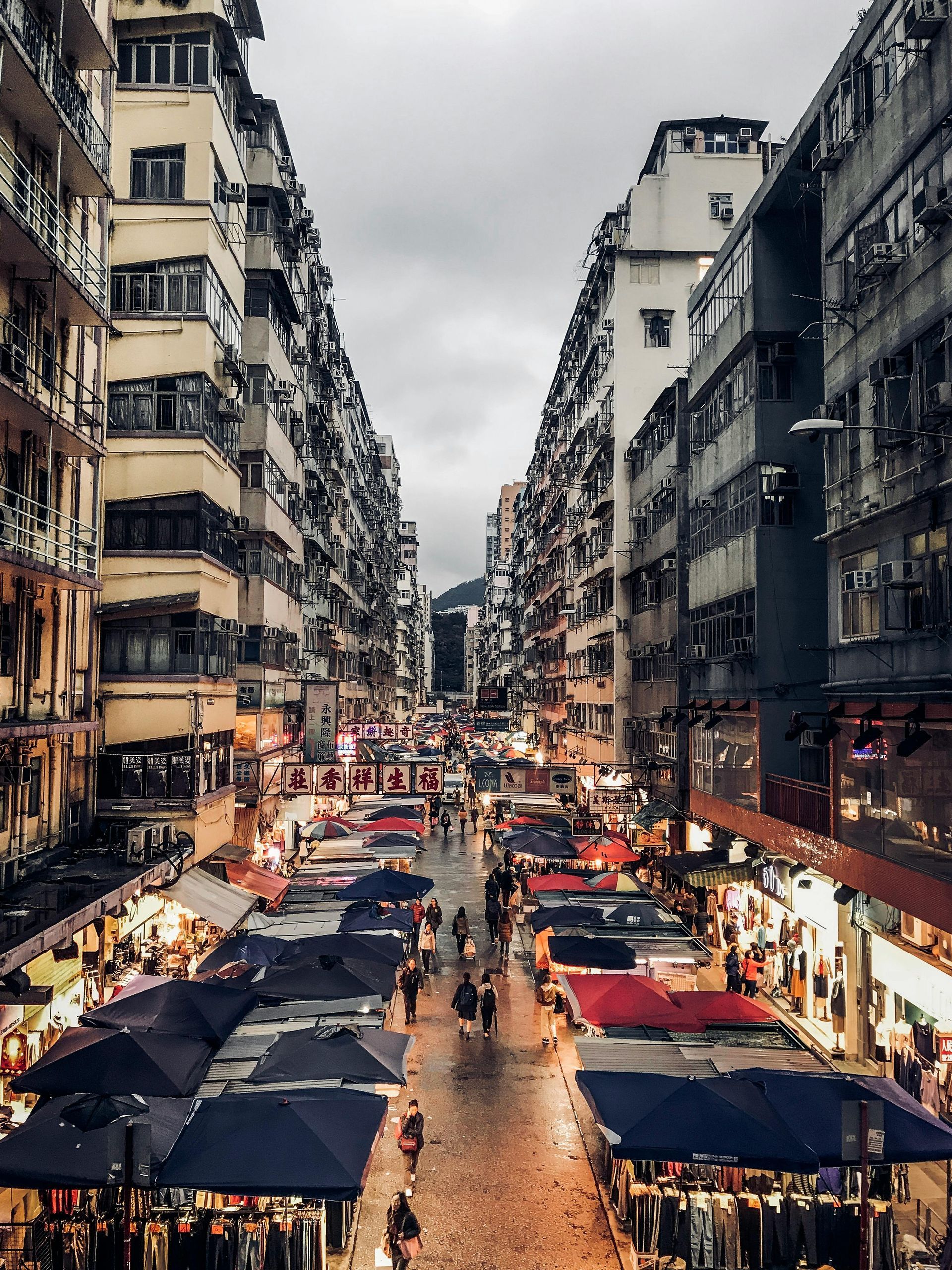阴天时，香港一条狭窄而熙熙攘攘的街市，两旁林立着高楼大厦和摊位。