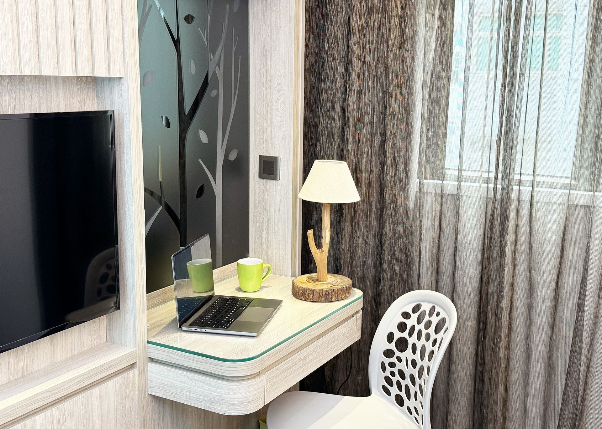 Desk with laptop, lamp, and chair near window with textured curtains and TV screen.