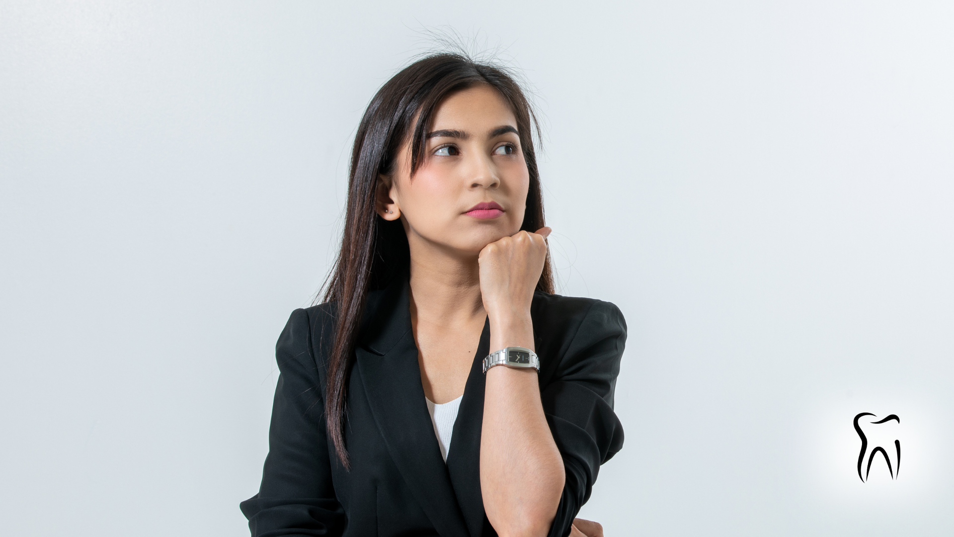 Woman in black blazer thoughtfully touching chin, looking up, white background.