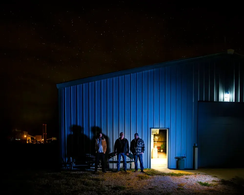 Three men stand outside a blue building under a starry night sky. Light shines from the doorway.
