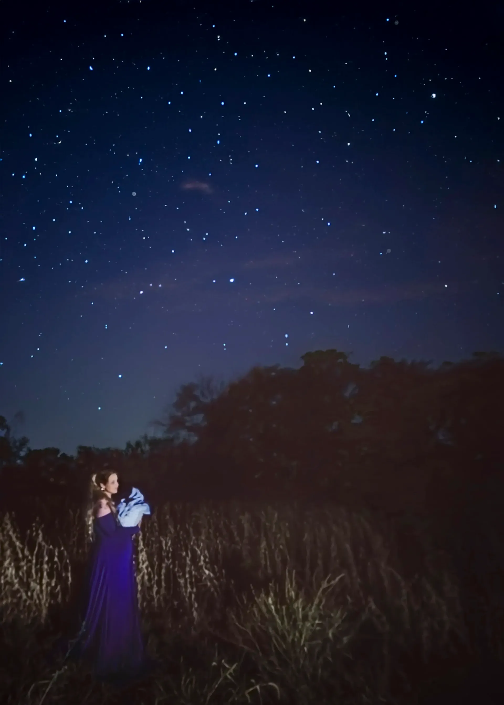 Woman in blue dress holding baby, lit against a starry night sky in a field.
