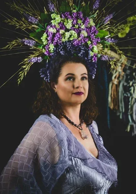 Woman in lavender gown and floral headdress, gazing towards the camera.
